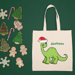 Cute Christmas Dinosaur Custom Green Kids Name トートバッグ<br><div class="desc">Awesome green dinosaur looking very cute in a holiday red Santa hat with a little smile. Adorable custom kids tote bag gift for your little boy or girl.</div>